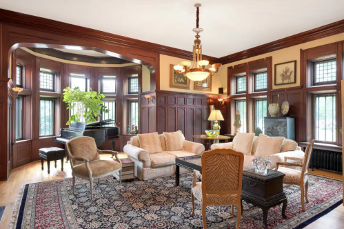 Elegant living room with wooden paneling, large windows, a grand piano, and classic furniture on a patterned rug.
