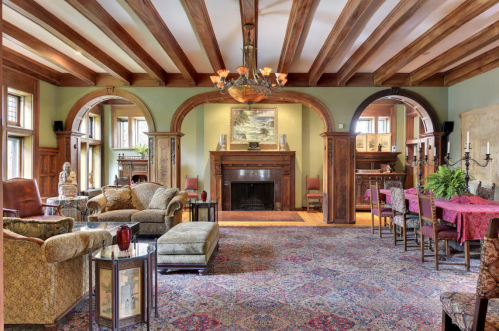 Elegant living room with wooden beams, a fireplace, plush seating, and a dining area, adorned with rich decor and rugs.