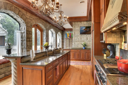Spacious kitchen with stone walls, wooden cabinets, a chandelier, and large windows overlooking greenery.
