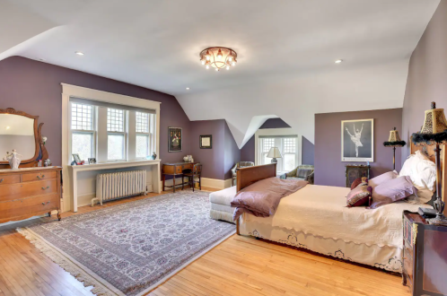 Spacious bedroom with purple walls, a large bed, vintage furniture, and a cozy seating area by the window.