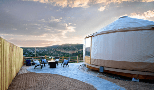 A cozy yurt with a fire pit and chairs, set against a scenic mountain view at sunset.