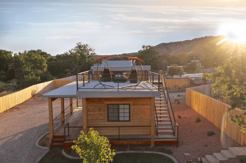 A modern wooden house with a rooftop swing, surrounded by trees and a gravel yard, under a sunset sky.