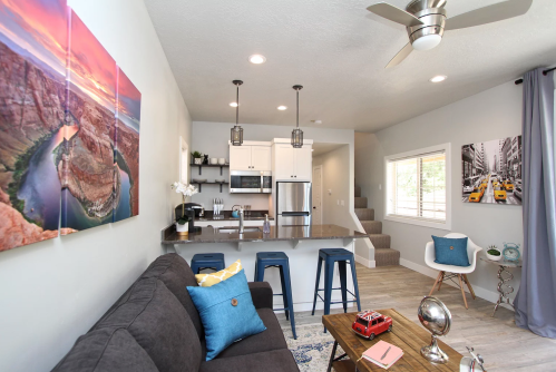Modern living room with a gray sofa, blue stools, and a kitchen area, featuring artwork and natural light.