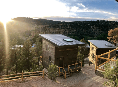 Two modern wooden cabins on a hillside, surrounded by trees, with a sunset illuminating the landscape.
