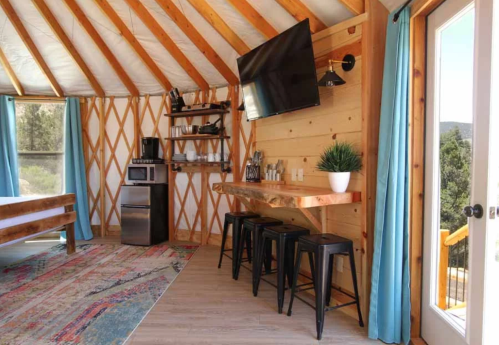 Cozy interior of a yurt featuring a wooden table, stools, kitchenette, and large windows with blue curtains.