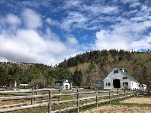 A scenic farm with a large barn, fenced areas, and lush green hills under a partly cloudy sky.