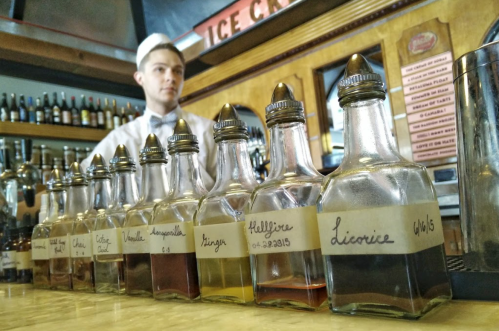 A barista stands behind a counter with various glass bottles of flavored syrups lined up in front.