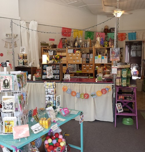A colorful craft shop interior with handmade items, greeting cards, and decorative banners. Bright and inviting atmosphere.