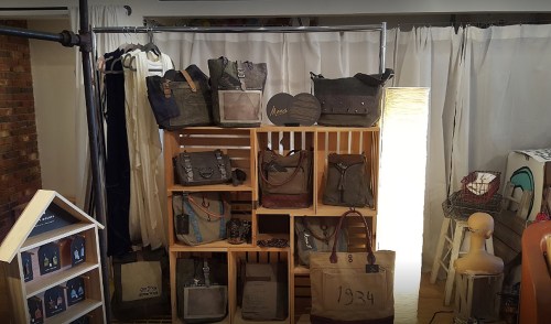 Display of various bags on wooden shelves, with a soft light and white curtains in the background.