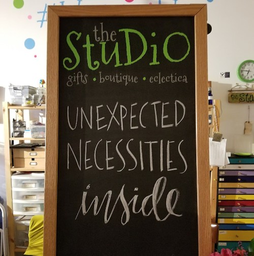 A chalkboard sign for "the Studio" boutique, advertising "Unexpected Necessities Inside." Colorful and inviting design.