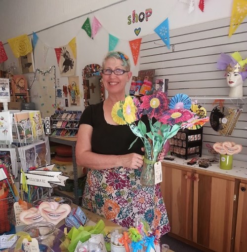 A smiling woman holds a vase of colorful paper flowers in a vibrant craft shop filled with decorations and art supplies.