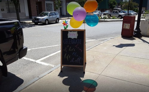 A sidewalk sign with colorful balloons, promoting a celebration in a town square.
