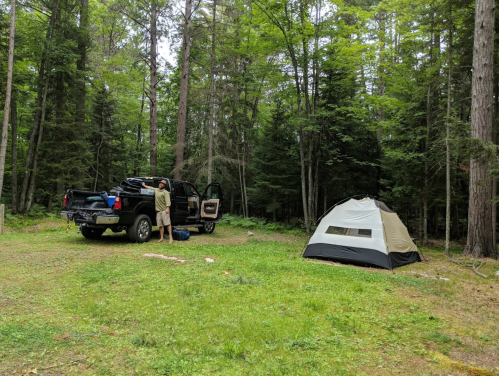 A person sets up a tent near a black truck in a wooded campsite surrounded by trees.