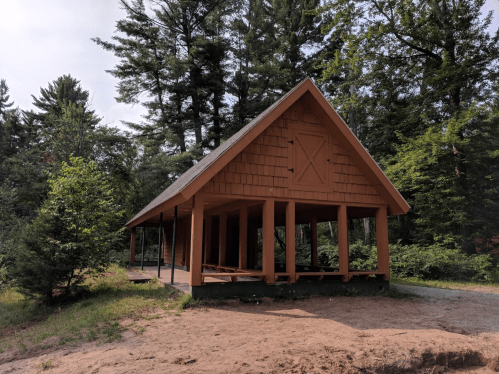 A wooden shelter with a sloped roof, surrounded by trees and greenery, set in a sandy area.