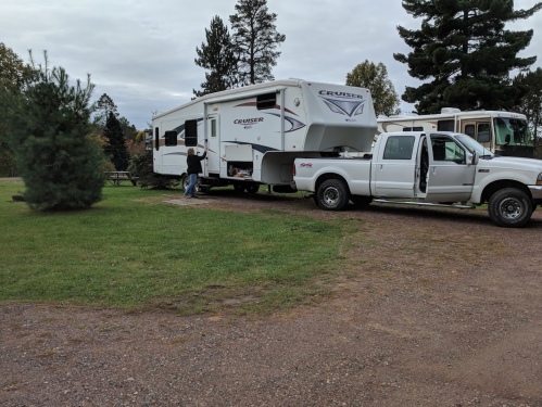 A white pickup truck is towing a large RV in a grassy area with trees and a cloudy sky in the background.