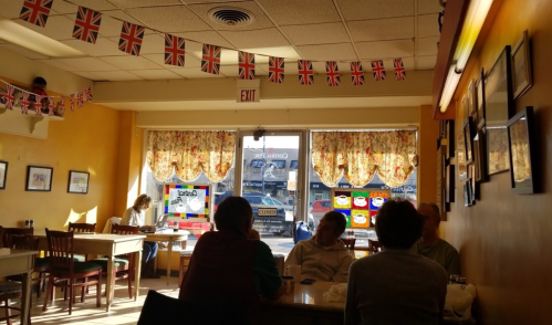 A cozy café with British flags, sunlight streaming in, and patrons chatting at tables near a window.