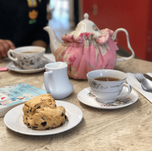 A cozy tea setting with a teapot, teacup, scone, cream pitcher, and a book on a marble table.