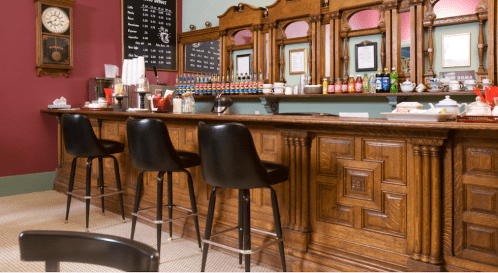 A vintage bar with wooden paneling, three black stools, and a menu board in a cozy, colorful setting.
