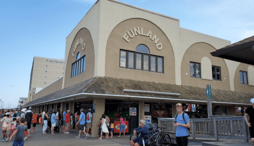 A busy boardwalk scene featuring the Funland building, with people gathered outside and a clear blue sky above.