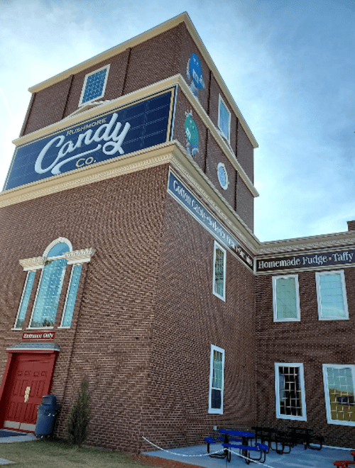 Exterior of a brick building featuring the "Ruby's More Candy Co." sign, with a red door and benches outside.
