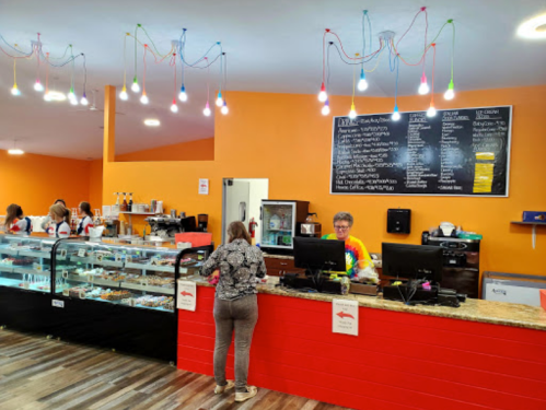 A colorful café interior with a counter, menu board, and customers enjoying treats. Bright lights hang from the ceiling.
