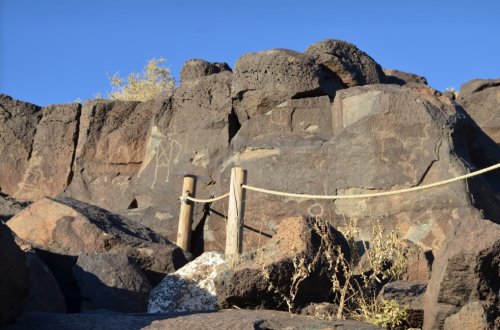Rock formations with petroglyphs and wooden posts tied with rope against a clear blue sky.