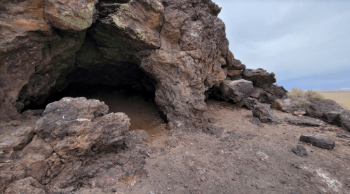 A rocky cave entrance surrounded by rugged terrain under a cloudy sky.