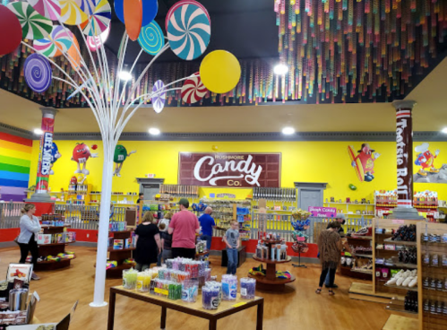 Brightly colored candy store interior with shelves of sweets, a rainbow wall, and whimsical decor. Shoppers browsing.