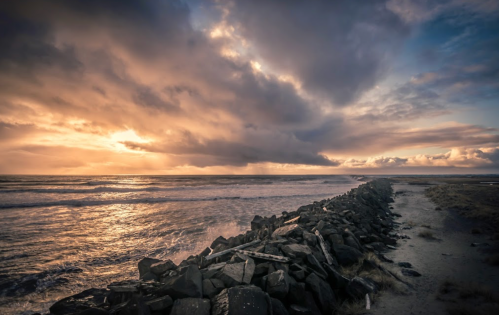 A rocky jetty extends into a turbulent sea under a dramatic sky at sunset, with clouds illuminated by warm light.