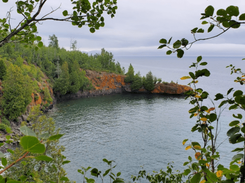 A serene coastal view with rocky cliffs, lush greenery, and calm waters under a cloudy sky.