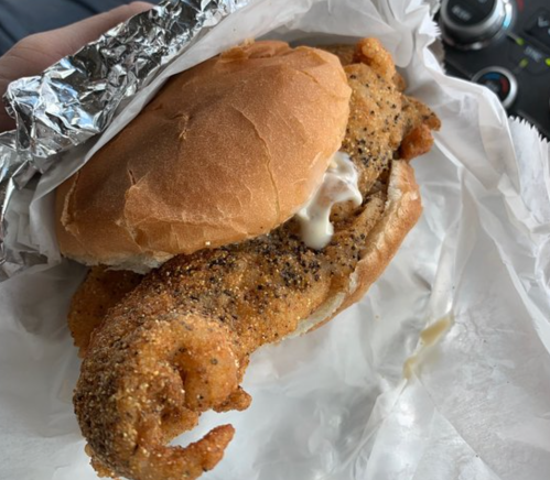 A fried fish sandwich with a crispy fillet and mayonnaise, wrapped in foil and placed on white paper.
