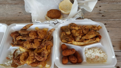 Two takeout containers with fried seafood, including shrimp, crab, fish, fries, coleslaw, and a biscuit.