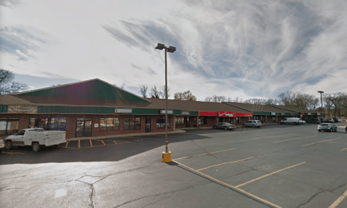 A shopping plaza with several storefronts, a red awning, and a cloudy sky above. Empty parking lot in front.
