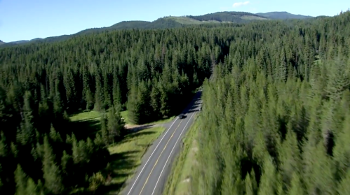 Aerial view of a winding road through dense green forests and mountains under a clear blue sky.