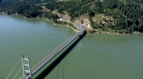 Aerial view of a suspension bridge spanning a green river, surrounded by lush forests and rocky terrain.