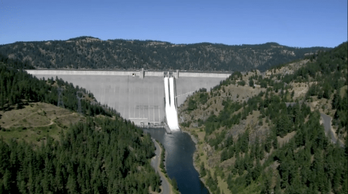 A large dam with water cascading down, surrounded by green hills and a clear blue sky.
