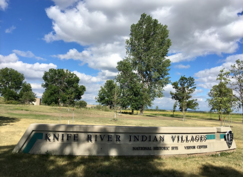 Sign for Knife River Indian Villages National Historic Site Visitor Center, surrounded by trees and a blue sky.