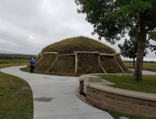 A woman stands near a large, grassy earth lodge with wooden supports, surrounded by a paved path and trees.