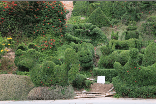 A lush garden featuring creatively shaped topiary bushes resembling various animals and forms, surrounded by colorful flowers.