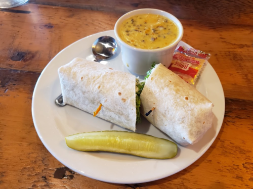 A plate with two wrapped burritos, a bowl of soup, a pickle, and a packet of sauce on a wooden table.