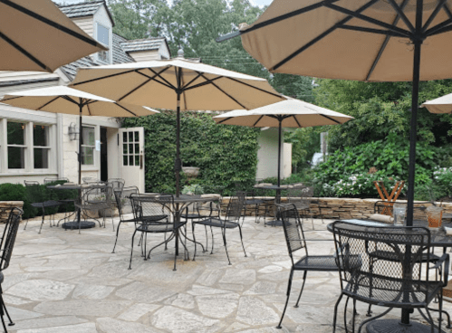 Outdoor dining area with black metal tables and chairs, shaded by large umbrellas, surrounded by greenery.