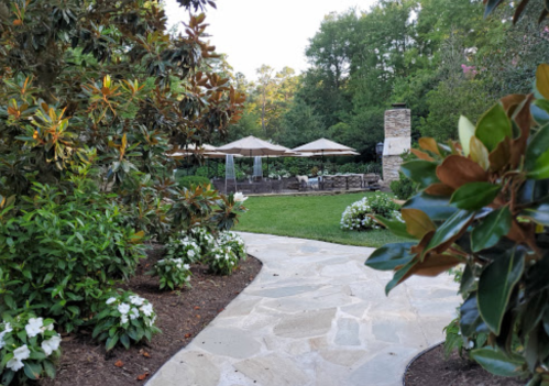 A stone pathway leads through lush greenery to a patio with umbrellas and seating in a serene garden setting.