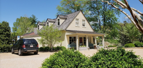 A charming light-colored house with a porch, surrounded by greenery and a gravel driveway, featuring a parked black SUV.
