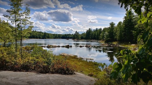 A serene lake surrounded by trees and rocks, reflecting clouds and blue sky in a peaceful natural setting.