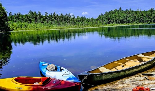 Three colorful kayaks are docked by a calm lake surrounded by lush green trees under a clear blue sky.
