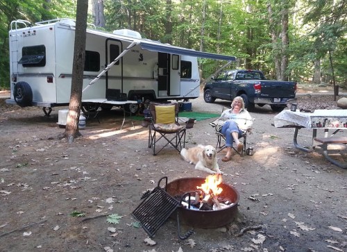 A woman relaxes by a campfire with a dog, near a camper and a pickup truck in a wooded campsite.