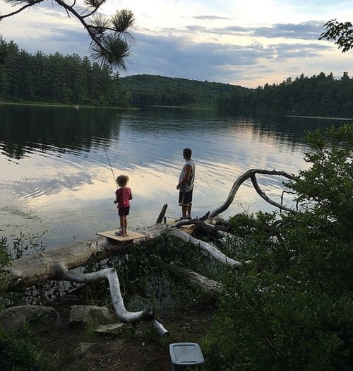 Two children fish from a log by a calm lake, surrounded by trees and a cloudy sky.