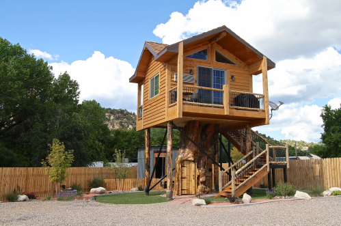 A wooden treehouse on stilts, featuring a balcony and surrounded by a fenced yard with trees and landscaping.