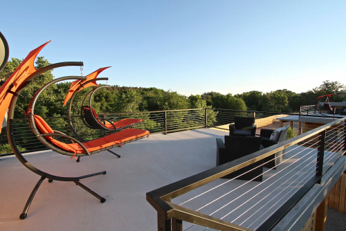 A spacious balcony with orange hanging chairs and a seating area, surrounded by lush green trees under a clear blue sky.