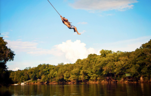 A person swings from a rope over a calm river, surrounded by lush green trees and a clear blue sky.